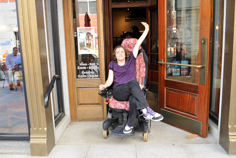 Woman in wheelchair rolling out of newly accessible cafe with her hand up in celebratory gesture