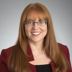 Professional headshot of Judith Bomster. Gray background. Long reddish hair, glasses, smile, maroon suit coat, black shirt, pearl necklace.