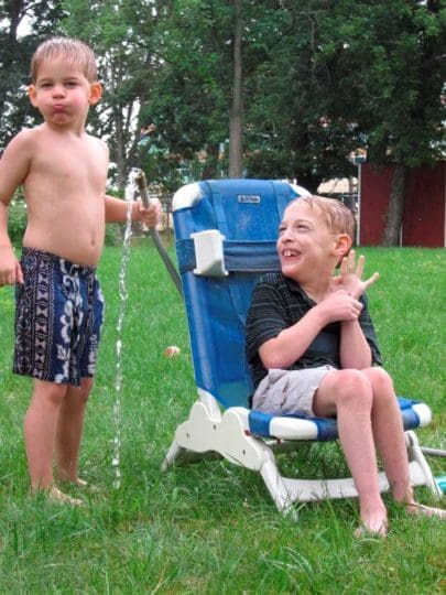 Two boys playing in sprinkler in yard. One boy sitting in a waterproof chair.
