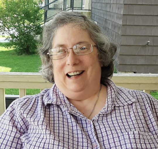 Ellen smiling while sitting outside. She has short hair with glasses and is wearing a purple checkered shirt. 