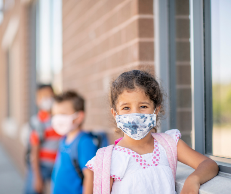 Young student with mask leans against brick wall of school. Other students are visible socially distant behind her.