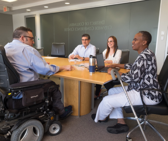 Group of employees with a variety of skin tones sit at a conference table. A person using a wheelchair speaks with a person using a cane.
