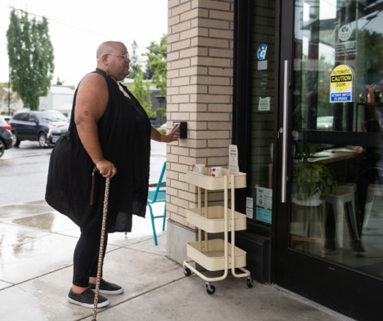 Person with medium-dark skin tone in black dress and cane presses button for automatic door to open at a store entrance
