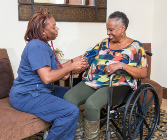 Healthcare provider in blue scrubs checks the vitals on a person sitting in a wheelchair. Both people are people of color.