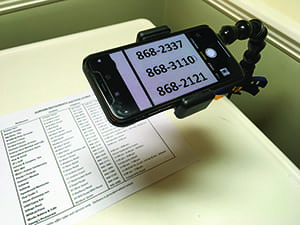 A smart phone screen is visible while sitting in a black hands-free cell phone holder. The holder is attached to a table. A piece of paper on the table has a lot of indecipherable content. The phone’s screen is magnifying some of the numbers on the page below it.