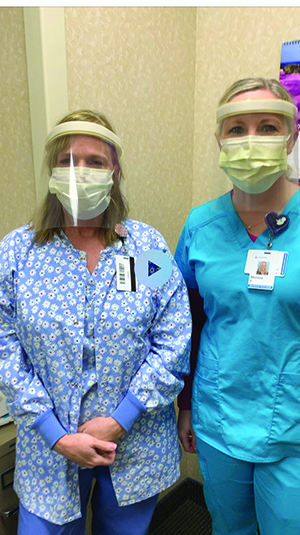 Two health care workers wearing masks and scrubs model face shields.