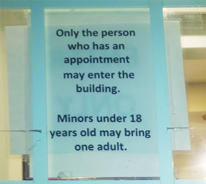A sign is taped to a glass door reads Only the person who has the appointment may enter the building. Minors under 18 years old may bring one adult.