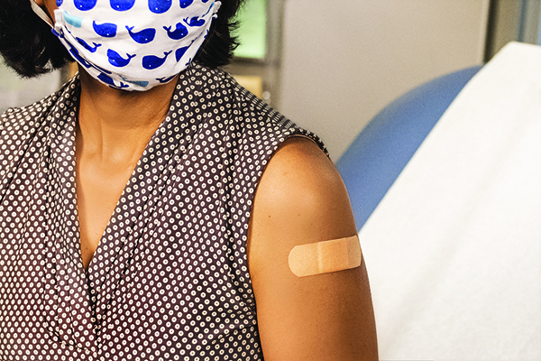 Person wearing mask sits in a medical office. A band-aid is visible on their arm.