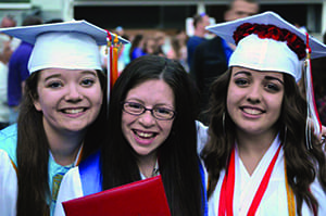 Three students posing at a high school graduation, they smile excitedly as one student holds up their diploma to the camera.