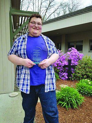 Smiling person in purple shirt and jeans holding an award.
