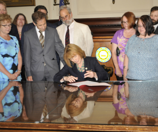 Former Governor Maggie Hassan signs the CMHA while sitting at large desk surrounded by advocates. 
