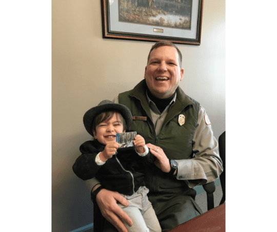 Young child wearing a fedora sits in lap of NH park ranger. Both laugh while child proudly holds his new park pass.