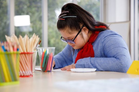 Student with medium light skin tone, black hair, and blue shirt with red scarf focuses on school work at a light colored table. Containers of pencils and crayons are nearby.