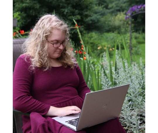 Sophie with light skin tone, short blond hair, glasses, and deep red top sitting outside working on her laptop.