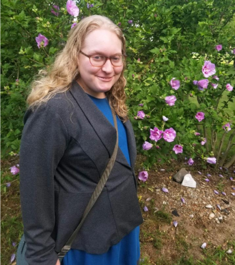 Sophie with light skin tone, short blond hair, glasses, and wearing a suit coat and work bag standing in front of some rose bushes