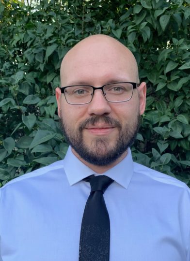 Headshot of Ryan who is bald with closely trimmed facial hair and wears glasses and a blue button down shirt and dark tie.
