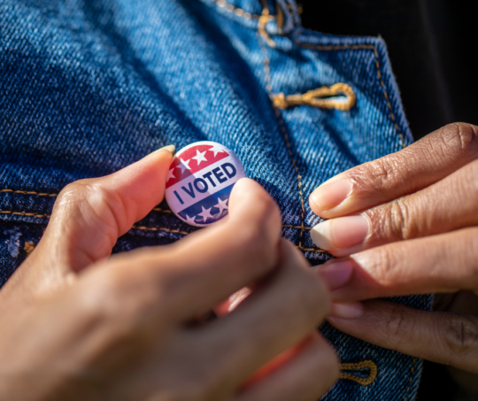 Close up of hand as person pins an 'I Voted' button on to their jean jacket.