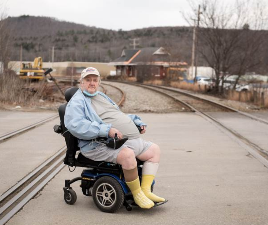 James wears a white ball cap, blue shirt and yellow socks and sits in his wheelchair and looks at camera. Rail road tracks are on both side of him.