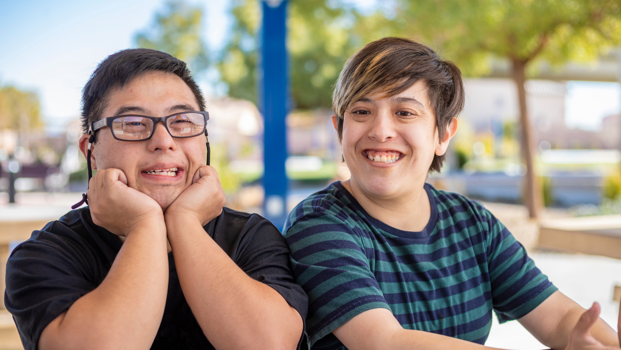 Two young adults with disabilities sit outside together smiling. The person on the left has medium skin tone, short dark hair, a black t-shirt, and is wearing glasses. The person on the right has light skin tone, short brown hair, and is wearing a blue striped shirt.