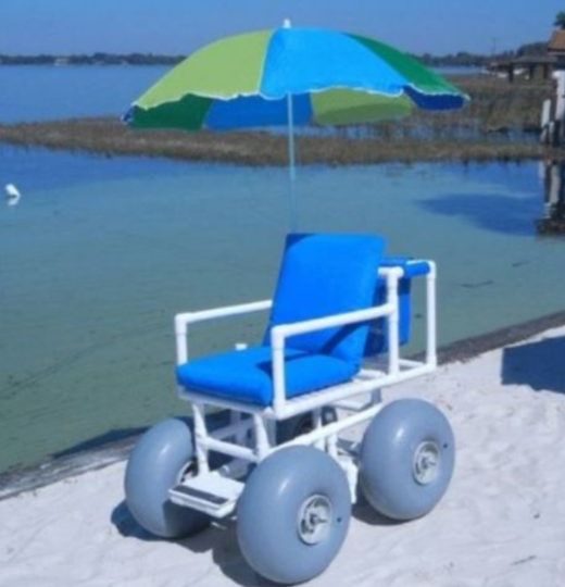 Blue and white beach wheelchair with large gray wheels and sun umbrella on sandy beach with water in the background.