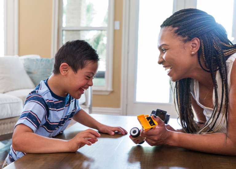 A parent and child with medium dark skin tone sit around a coffee table laughing. The child appears to have disabilities and the parent holds a yellow plastic toy.