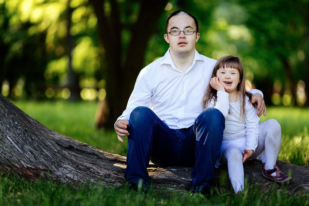 Parent and child, both with disabilities, sit together on tree root surrounded by bright forest greenery.
