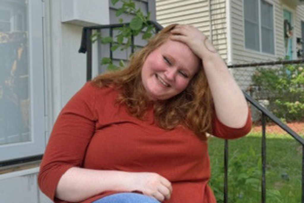 A beaming young woman sits on a front stoop and runs her hand through her reddish blonde hair.